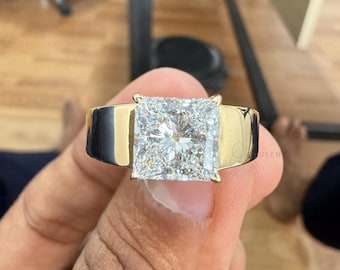 May include: A wide gold band ring featuring a large, square-cut, faceted clear gemstone set in the centre. The metal band has a polished, reflective surface that contrasts with the brilliant, multi-faceted sparkle of the central stone.