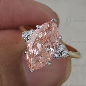 May include: A ring featuring a large, marquise-cut peach-coloured gemstone set in a metal band. The central stone is flanked on each side by a cluster of three small, round, clear white stones held in place by silver-toned prongs.