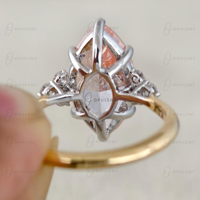 May include: A ring featuring a large marquise-cut peach-coloured gemstone set in a silver-toned prong setting, accented by smaller clear stones on each side. The ring has a smooth gold-toned band and is marked with the text OPULENT throughout the image.
