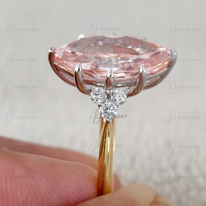 May include: A ring featuring a large, marquise-cut peach-coloured gemstone set in a silver-toned metal prong setting. The band is gold-toned, and the setting is accented with three small, round, clear white diamonds on each side of the centre stone.