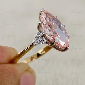 May include: A ring featuring a large marquise-cut peach-coloured gemstone set in a silver-toned metal prong setting. The band is made of gold-toned metal and is accented with three small round-cut clear stones on each side of the centre stone.