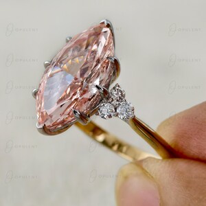 May include: A ring featuring a large marquise-cut peach-coloured gemstone set in a silver-toned metal prong setting. The band is made of yellow gold and includes a cluster of three small, round, clear white accent stones on each side.