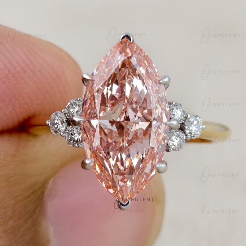 May include: A marquise-cut peach-coloured centre stone set in a metal ring, featuring a cluster of three small round white accent stones on each side of the main setting. The ring band appears to be a metallic gold tone with silver-toned prongs.