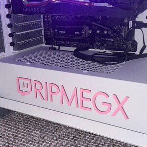 以下が含まれることがあります： ピンク色の「RIPMEGX」デカールが付いた白いコンピューターケース。デカールにはTwitchのロゴが含まれています。ケースはメッシュトップで、GIGABYTEグラフィックカードなど、内部コンポーネントが見えます。
