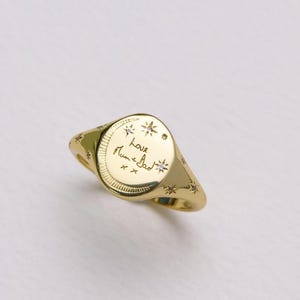 Puede incluir: Un anillo de sello dorado con una cara ovalada. El anillo presenta un diseño grabado con estrellas y escritura. El anillo está sobre un fondo blanco. El anillo tiene aproximadamente 1,9 cm de diámetro.