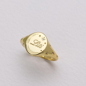 Puede incluir: Un anillo de sello dorado con el nombre "Lili" grabado en una cara circular. El anillo presenta detalles de estrellas y está sobre un fondo blanco.