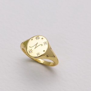 Può includere: Un anello con sigillo d'oro con una faccia ovale. L'anello presenta una lettera "N" incisa e disegni a forma di stella attorno alla faccia. L'anello è posto su uno sfondo bianco.