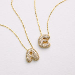 May include: Two gold necklaces with pendant letters, "A" and "E", encrusted with small, clear gemstones. The pendants are attached to delicate gold chains. The necklaces are displayed on a white surface.