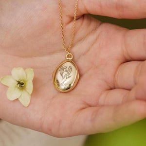 May include: A gold-colored necklace with a pendant featuring a floral design is held in an open hand, next to a small white flower. The background is a soft green and cream color.