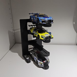 Puede incluir: Un expositor negro de tres niveles que sostiene tres coches de carreras en miniatura. Los coches son azules, amarillos y plateados, cada uno con calcomanías de carreras detalladas. El expositor está diseñado para exhibir los modelos de coches.