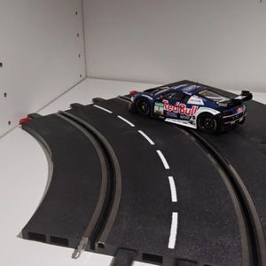 Könnte beinhalten: Ein blaues, weißes und rotes Rennwagenmodell mit dem Red Bull-Logo steht auf einer schwarzen Autorennbahn. Die Bahn hat weiße Fahrbahnmarkierungen und silberne Schienen. Das Auto hat einen Spoiler und ist auf der Bahn abgewinkelt.