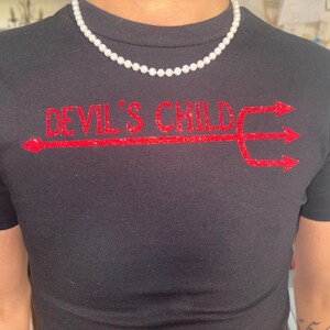 May include: Black crew neck t-shirt with the text "DEVIL'S CHILD" in red glitter lettering and a red glitter trident design. A white pearl necklace is worn around the neck. The shirt is a close-up shot.