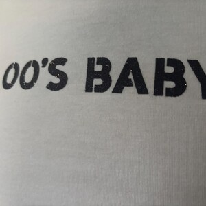 May include: White t-shirt with the text "00'S BABY" in black, glittery letters. The shirt is made of a soft material and is suitable for casual wear. The text is centered on the chest.