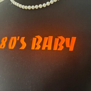 May include: Black t-shirt with the orange text "80'S BABY". A white pearl necklace is visible at the neckline. The shirt is a casual wear item, perfect for a retro or vintage style.
