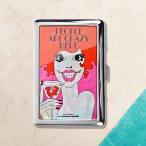 May include: A silver metal cigarette case featuring a retro illustration. The artwork depicts a woman with red hair and a martini glass, with the text "PEOPLE ARE CRAZY HERE." The case also has the text "By the Author of DO YOU SLEEP IN THE NUDE?"