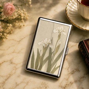 May include: A rectangular metal cigarette case with a polished silver-tone frame and a central panel featuring a botanical illustration of white iris flowers with green stems and leaves against a light, textured, off-white background.