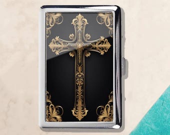 Puede incluir: Un estuche de cigarrillos de metal plateado con un diseño de cruz dorada. La cruz es ornamentada con filigranas detalladas sobre un fondo negro. El estuche tiene una superficie brillante y reflectante.