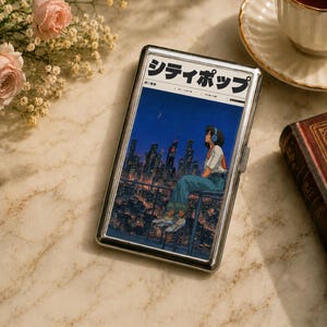 May include: A rectangular silver metal cigarette case featuring an anime-style illustration of a person wearing headphones, sitting on a balcony overlooking a city skyline at night. The top of the case displays the Japanese text シティポップ.