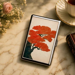 May include: A rectangular metal cigarette case with a polished silver frame and a flat top surface featuring a printed illustration of a dark tree trunk with vibrant orange-red foliage against a cream-coloured background.
