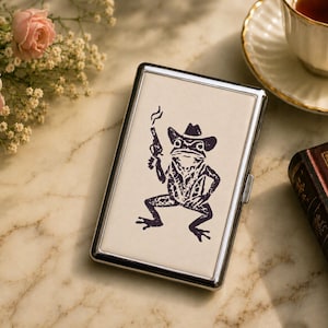 May include: A rectangular silver metal cigarette case featuring a cream-colored front panel. The panel displays a dark purple graphic illustration of a frog wearing a cowboy hat, holding a smoking pistol, and standing in a confident pose.