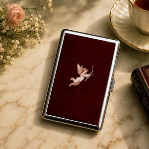May include: A rectangular metal cigarette case with a polished silver-tone frame and a deep burgundy velvet inlay on the front. The center of the velvet panel features a small, detailed white illustration of a winged cherub holding a bow and arrow.