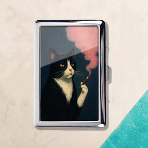 May include: A silver cigarette case featuring a black and white cat with yellow eyes, holding a lit cigar. The cat is surrounded by pink smoke against a dark blue background. The case has a shiny, metallic finish.