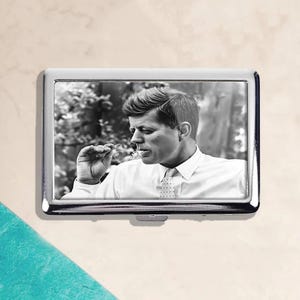 May include: A silver cigarette case featuring a black and white image of a man in a white shirt and tie. The image is a portrait of John F. Kennedy. The case has a rectangular shape with rounded corners.