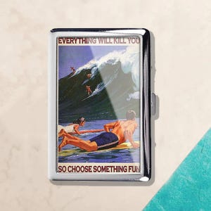 May include: A silver cigarette case with a surfing-themed graphic. The image features surfers riding a large wave, with the text "EVERYTHING WILL KILL YOU SO CHOOSE SOMETHING FUN". The case has a shiny, metallic finish.