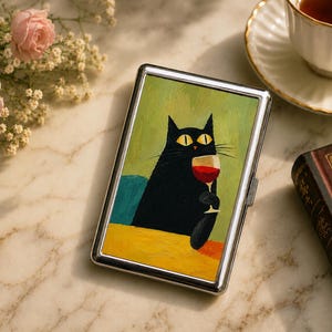 May include: A rectangular metal case featuring a printed illustration of a black cat with yellow eyes holding a glass of red wine. The cat is positioned against a green background with a yellow foreground surface. The case has a polished silver frame.