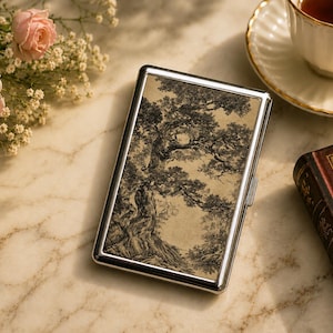 May include: A rectangular metal cigarette case with a polished silver finish and a hinged lid. The front panel features a detailed black and white ink illustration of a gnarled, ancient tree set against a light beige, parchment-like background.