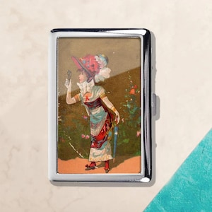 May include: A silver cigarette case featuring a vintage illustration of a woman in a colorful dress and hat, holding a small object. The artwork has a classic, antique aesthetic, with a garden backdrop.