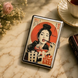 May include: A rectangular metal cigarette case featuring a graphic illustration of a person with a dark bun hairstyle eating from a red bowl with chopsticks. The design includes a large red circle, cream background, and black Japanese text characters.