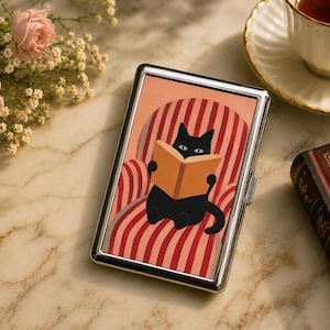 May include: A rectangular silver metal cigarette case featuring a graphic illustration of a black cat with blue eyes reading an open book while sitting in a red and white striped armchair. The case has a polished metallic frame and a hinged lid.