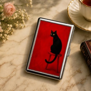 May include: A rectangular metal cigarette case featuring a vibrant red background with a graphic illustration of a black cat with yellow eyes sitting on a small red stool. The case has a polished silver-tone frame and a hinged opening mechanism.