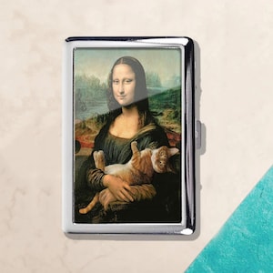 May include: A silver metal cigarette case featuring a humorous image of the Mona Lisa holding a ginger cat. The artwork is a parody of the famous painting, with the cat replacing the original subject. The case has a rectangular shape and a shiny finish.