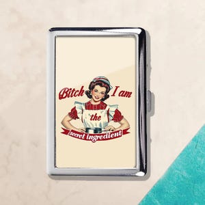 May include: A silver metal cigarette case featuring a vintage illustration of a woman in a red and white apron. The text reads "Bitch I am the secret ingredient" in red script.
