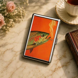 May include: A rectangular metal cigarette case featuring a vibrant orange background with a detailed illustration of a rainbow trout with green and pink scales, holding a lit cigarette in its mouth with a trail of smoke rising upward.