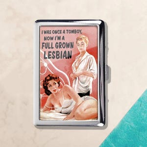 May include: A silver cigarette case with a retro-style image. The image features two women in vintage attire with the text "I WAS ONCE A TOMBOY, NOW I'M A FULL GROWN LESBIAN". The case has a shiny, metallic finish.