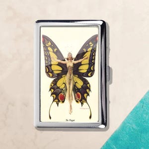 May include: A silver metal cigarette case featuring a vintage illustration of a woman with butterfly wings. The artwork is in yellow, black, and red, with the text "The Flapper" at the bottom. The case has a shiny, reflective surface.