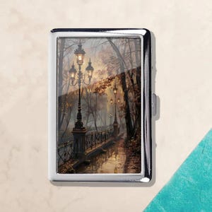 May include: A silver cigarette case featuring a scenic painting of a park path with lampposts. The artwork depicts a misty, autumn scene with a bridge, trees, and warm lighting, creating a serene atmosphere.