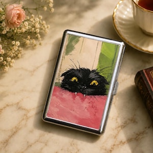 May include: A rectangular metal cigarette case featuring a printed illustration of a black cat with wide yellow eyes and visible white fangs peeking over a textured pink surface against a background of green and beige vertical panels.