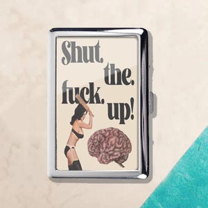 May include: A silver cigarette case with a graphic design. The design features the text "Shut the fuck up!" above an illustration of a woman hitting a brain with a baseball bat.