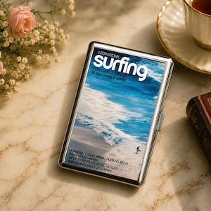 May include: A rectangular silver metal cigarette case featuring a glossy cover with an International Surfing magazine graphic. The image displays a blue ocean wave, a surfer, and text including "YOU CAN LEARN IT!" and "HAWAII, CALIFORNIA, PUERTO RICO."