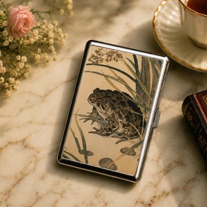May include: A rectangular metal cigarette case featuring a detailed illustration of a toad sitting among tall green grasses and small gray stones on a light beige background. The case has a polished silver-tone frame and a hinged side closure.