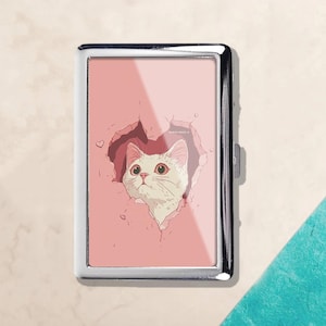 May include: A silver metal cigarette case featuring a pink background with a white cat illustration peeking through a heart-shaped tear. The cat has green eyes and a curious expression. A small heart is drawn on the left side.