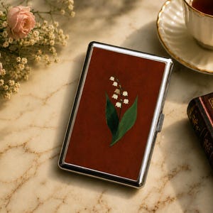 May include: A rectangular metal cigarette case with a polished silver frame and a deep rust-colored inlay on the front. The center of the inlay features a detailed illustration of a lily of the valley plant with white bell-shaped flowers and green leaves.