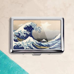 May include: A silver metal cigarette case featuring a print of Hokusai's "The Great Wave off Kanagawa." The artwork depicts a large ocean wave with boats and Mount Fuji in the background. The case has a rectangular shape with rounded corners.