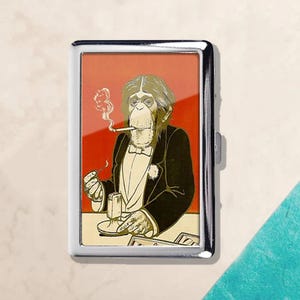 May include: A silver metal cigarette case featuring a chimpanzee illustration. The chimpanzee is depicted in a tuxedo, smoking a cigarette, with a drink on the table. The background is red.