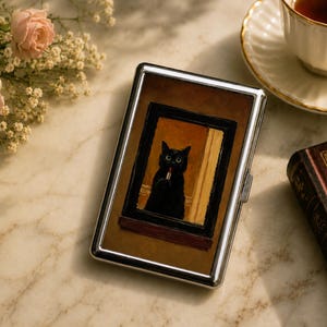 May include: A rectangular silver metal case featuring a printed image of a black cat inside a dark frame. The cat is depicted in a warm-toned room, holding a small red object. The case has a polished metallic border and a hinged closure mechanism.