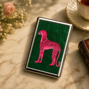 May include: A rectangular metal cigarette case featuring a vibrant green velvet inlay on the front cover. Centered on the green surface is a stylized illustration of a bright pink cheetah with black spots, depicted in a standing profile position.